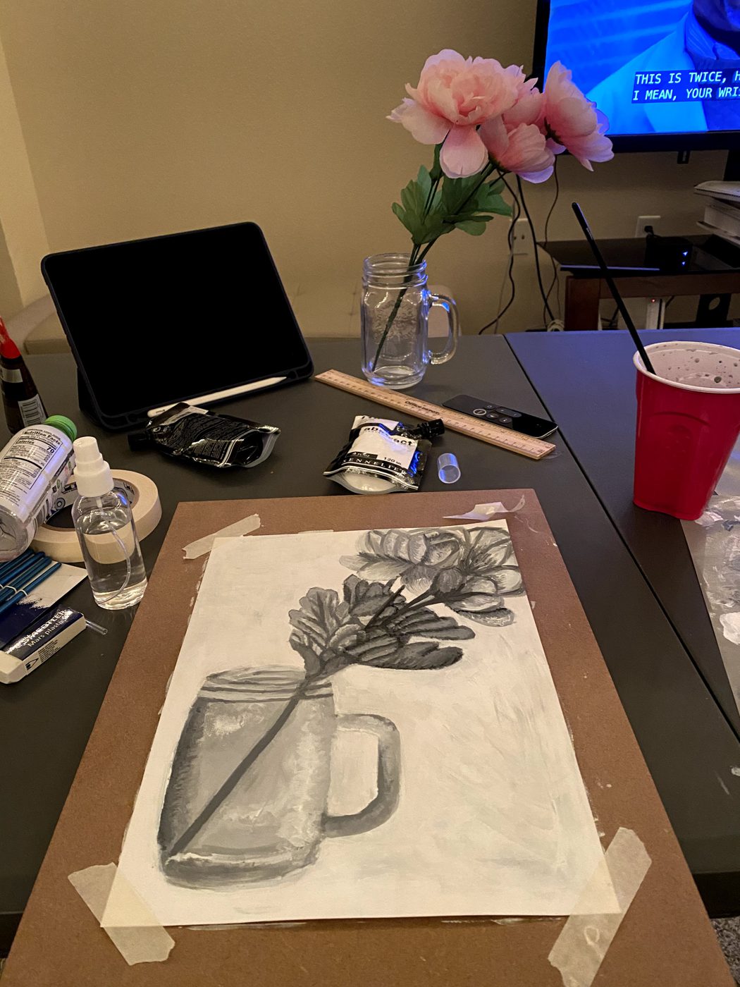 The gray painting taped to a board on a studio table, with the real pink flowers in a jar standing behind it.